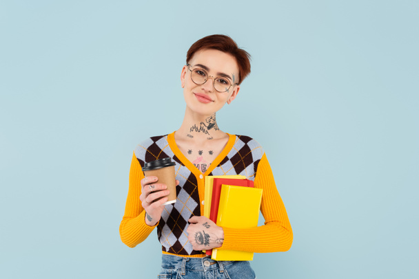 smiling tattooed student in glasses holding books and paper cup isolated on blue - Φωτογραφία, εικόνα