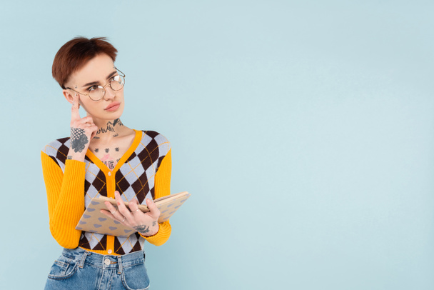 tattooed woman in glasses holding book in heart pattern cover and thinking isolated on blue - Zdjęcie, obraz