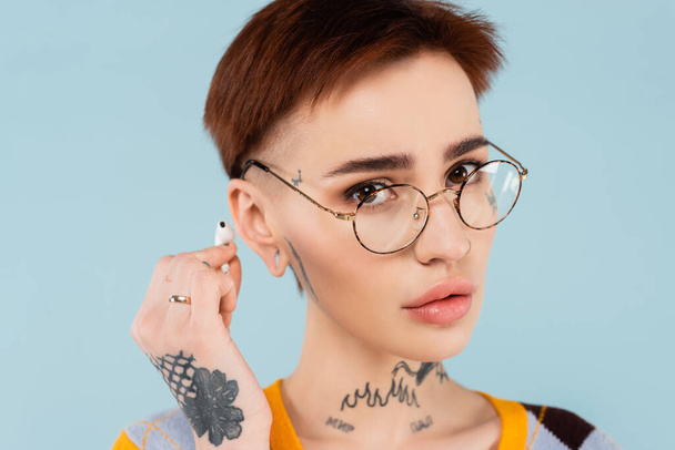 fiatal és tetovált nő szemüvegben tartja vezeték nélküli fülhallgató elszigetelt kék - Fotó, kép