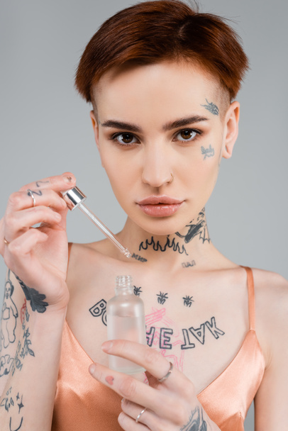 young tattooed woman holding bottle with serum and pipette isolated on grey  - Fotoğraf, Görsel