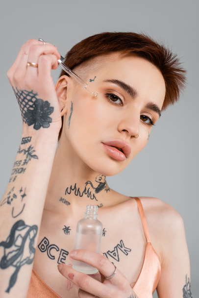 young tattooed woman holding bottle and pipette while applying serum on face isolated on grey  - Foto, Bild