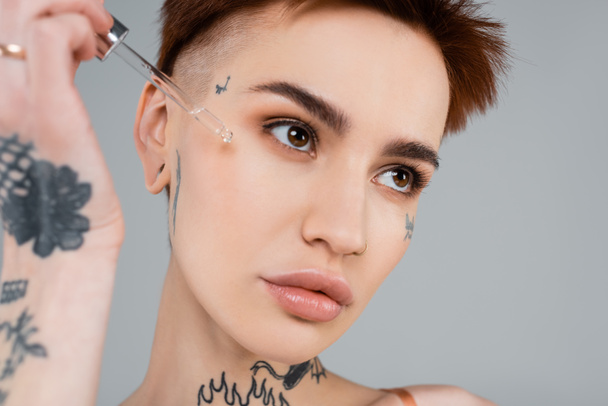 young tattooed woman applying serum with pipette isolated on grey  - Zdjęcie, obraz