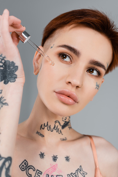 young tattooed woman applying serum on face with pipette isolated on grey  - Valokuva, kuva