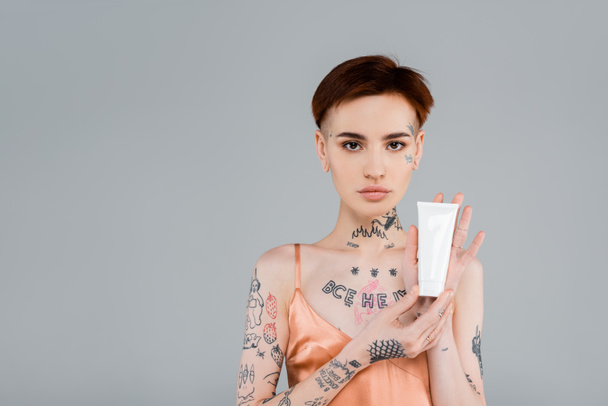 tattooed young woman holding tube with cosmetic product isolated on grey - Foto, Imagem