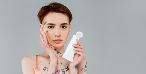 young woman with tattoos holding white tube and applying cream on face isolated on grey, banner - Фото, изображение