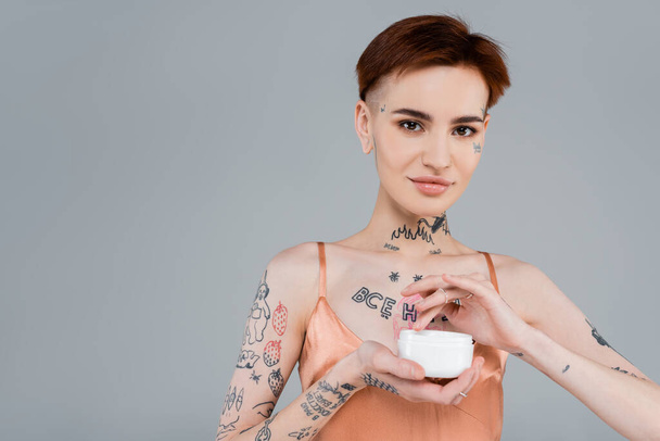 happy woman with tattoos and red hair holding white container with cosmetic product isolated on grey  - Foto, Imagen
