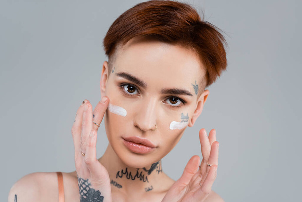 mujer joven tatuada aplicando crema hidratante en la cara aislada en gris  - Foto, imagen