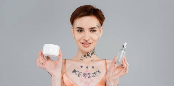 young and tattooed woman holding cosmetic products while smiling isolated on grey, banner  - Foto, Bild