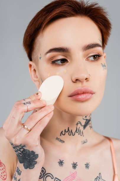tattooed woman applying makeup foundation with sponge isolated on grey - 写真・画像