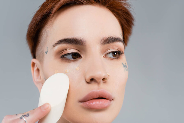 close up of tattooed woman applying makeup foundation on face with sponge isolated on grey - Photo, Image