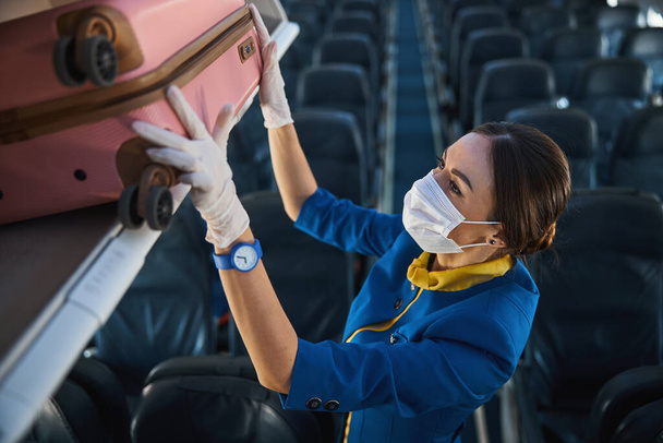 Airline stewardess carefully putting pink suitcase into luggage rack - Фото, изображение