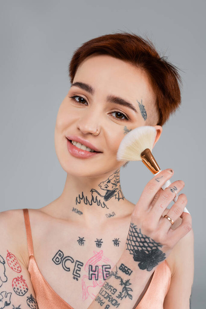 mulher tatuada alegre segurando escova cosmética ao aplicar pó de rosto isolado no cinza - Foto, Imagem