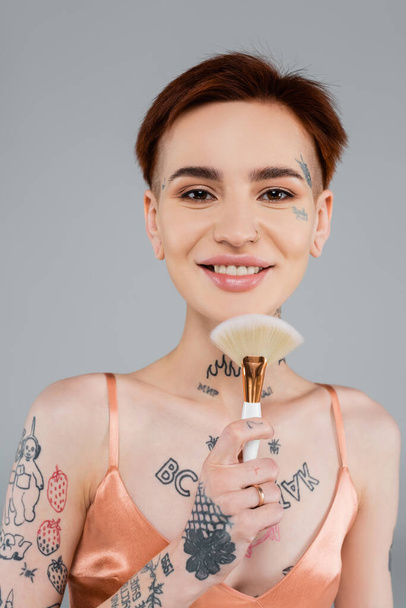 happy tattooed woman holding cosmetic brush isolated on grey - Photo, Image