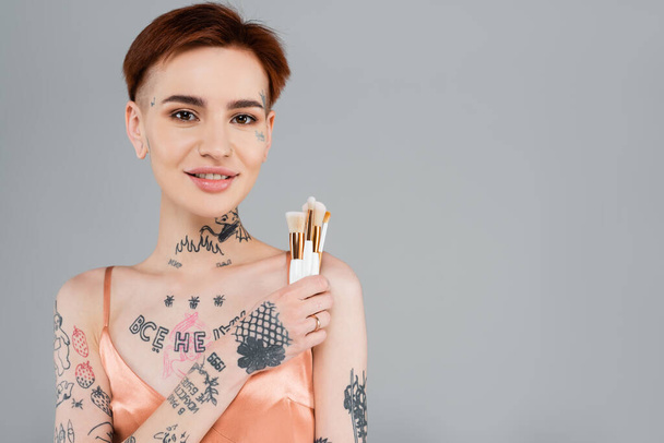 ευτυχισμένη και νεαρή γυναίκα με τατουάζ κρατώντας πινέλα καλλυντικών απομονωμένη σε γκρι - Φωτογραφία, εικόνα