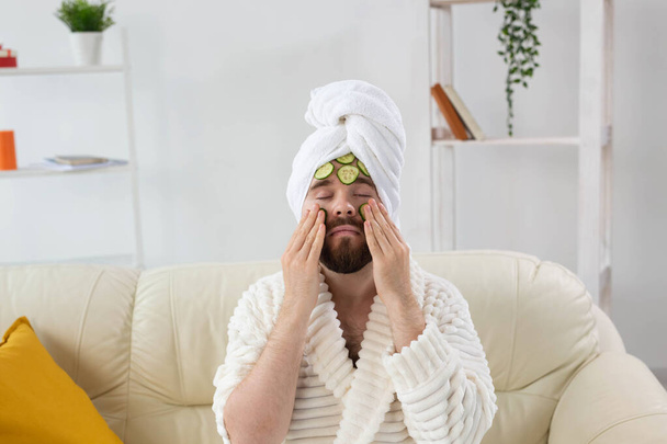 Baard man geniet met een cosmetisch masker op zijn gezicht gemaakt van plakjes komkommer. Mannen huidverzorging en spa thuis concept - Foto, afbeelding