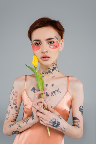 молодая и татуированная женщина в розовых повязках на глазу держит тюльпан изолированным на сером - Фото, изображение