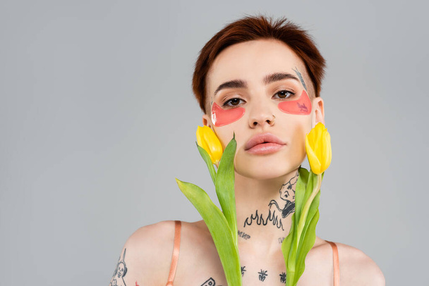 tetovaná žena v růžových páskách na očích držící žluté tulipány izolované na šedé - Fotografie, Obrázek