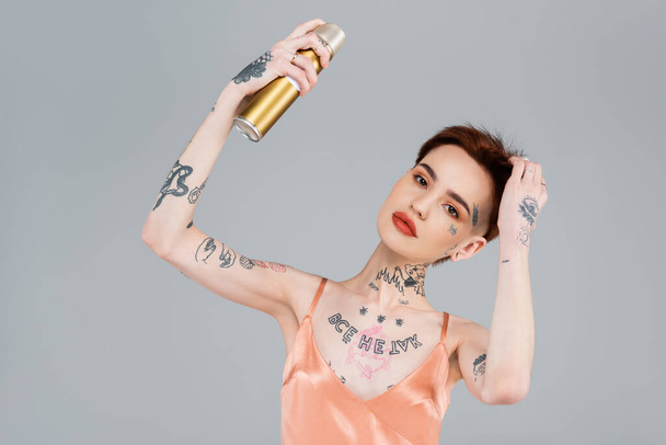 young tattooed woman with red lips holding hairspray isolated on grey - 写真・画像