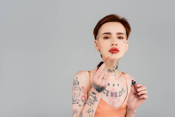mladá tetovaná žena s červenými rty nanášející lesk na rty izolovaný na šedou - Fotografie, Obrázek