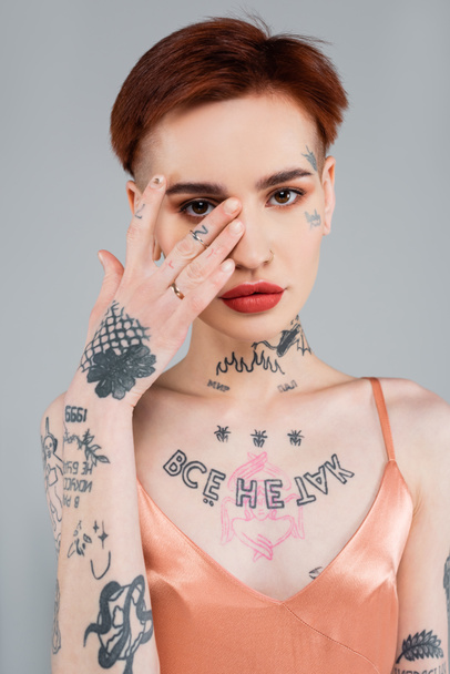 mujer joven tatuada con labios rojos cubriendo la cara con la mano aislada en gris  - Foto, Imagen