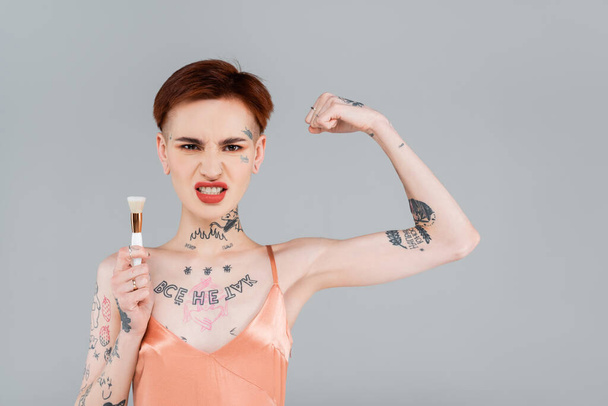 jeune femme tatouée avec des lèvres rouges montrant le muscle et tenant la brosse cosmétique isolée sur gris - Photo, image