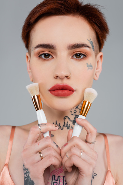 young tattooed woman with red lips holding cosmetic brushes near face isolated on grey - Fotoğraf, Görsel