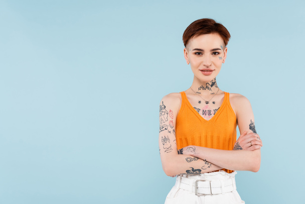 pleased and tattooed woman in orange knitted tank top standing with crossed arms isolated on blue  - Fotó, kép