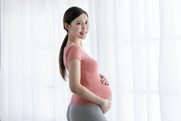 Side view of beautiful asian pregnant woman standing near window and smile at you in the living room - 写真・画像