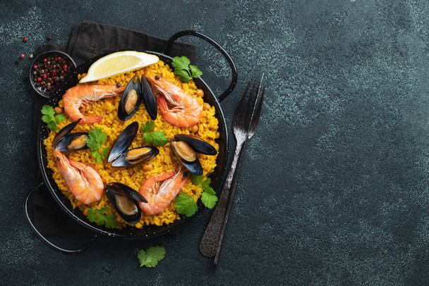 Traditionelle spanische Meeresfrüchte-Paella in der Pfanne mit Kichererbsen, Garnelen, Muscheln, Tintenfisch auf schwarzem Betonhintergrund. Draufsicht mit Kopierraum - Foto, Bild