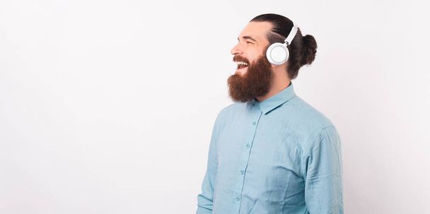 Portrait of smiling and laughing young man in casual with beard listening music at white wireless headphones. - Photo, Image