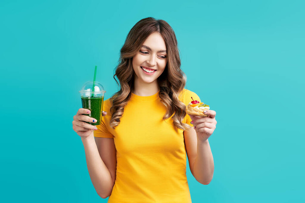 Happy woman choose unhealthy cake holding smoothie detox juice on blue background - Φωτογραφία, εικόνα