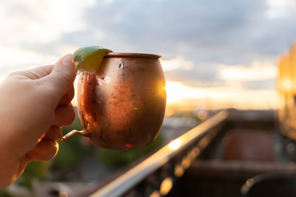 A moscow mule held up at a rooftop restaurant bar at sunset - Photo, Image
