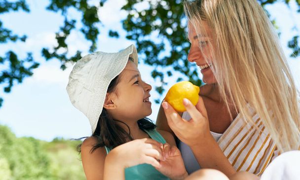Cheerful little girl looking to the lemon offered by her mother during sitting in the park. Happy young woman with her child having a cozy summer picnic with the family.  - Foto, imagen