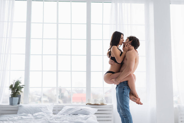Side view of sexy man holding girlfriend in lingerie in bedroom  - 写真・画像