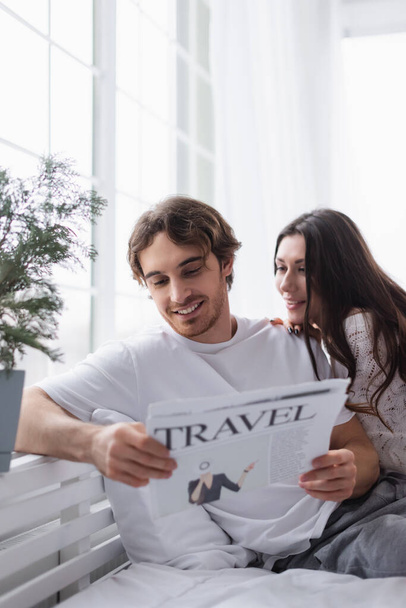 Smiling man reading travel newspaper near girlfriend on bed  - Zdjęcie, obraz