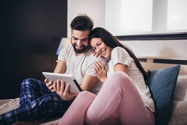 Jeune couple utilisant une tablette dans une chambre d'hôtel tout en étant couché sur le lit. - Photo, image