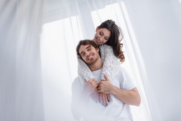 Young woman smiling while hugging boyfriend near window  - Fotografie, Obrázek