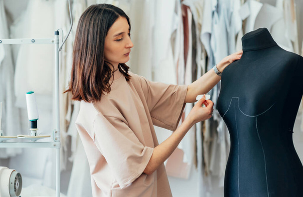 Side view shot of a fashion designer posing in her creative workshop. Young woman business owner working on the mannequin during preparing garment for a new collection in her creative office. - 写真・画像