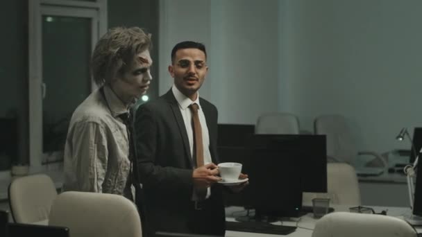 PAN schot van zakenman in pak en stropdas met een kop koffie en praten met zombie kantoormedewerker terwijl lopen met hem tussen bureaus - Video