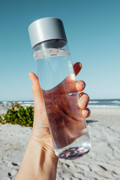 Mano femminile in possesso di bottiglia d'acqua potabile all'aperto sulla riva del mare. Concetto sanitario per il bilancio idrico. contenitore di acqua riciclabile. H2O - Foto, immagini