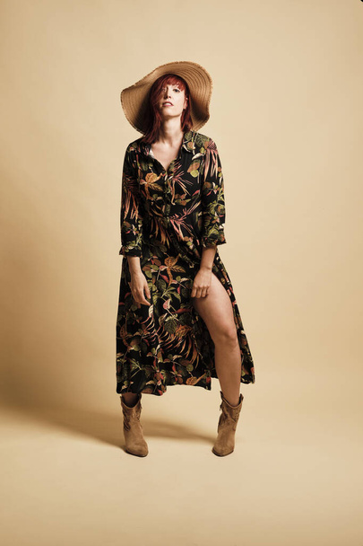 Pretty redhead woman smiling with a long floral dress and hat. Full-length, with boots and a yellow background. - Foto, Imagem