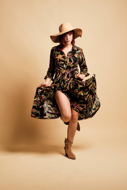 Pretty redhead woman smiling with a long floral dress and hat. Full-length, with boots and a yellow background. - Foto, Imagem