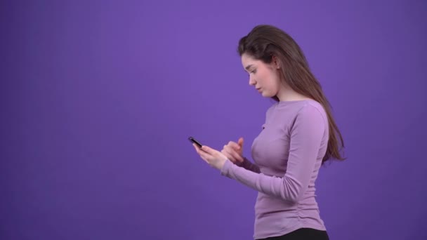 The charismatic girl, in profile, with the phone in hand, surfs the internet, looking for the location via GPS. Brunette isolated on a purple background, dressed in a purple sweater. Lifestyle concept - Video