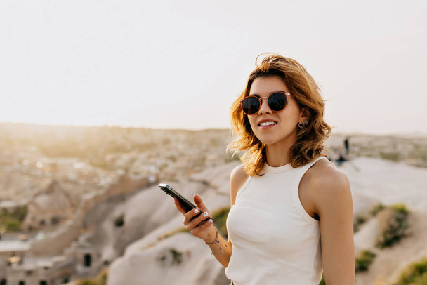 Stylish female tourist in sunglasses holding smartphone and walking among mountains with city view in sunlight . High quality photo - Photo, Image