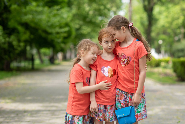 Three sisters stand in the center of the park hugging and saying something to each other in the ear, it's green on the street, the girls are dressed in bright orange dresses - Valokuva, kuva