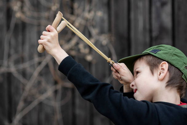A boy in a black jacket with a hood and a cap shoots from a slingshot, blurred background - Фото, зображення
