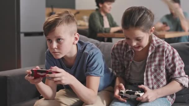 Średnie zbliżenie spowolnienie słodkie rodzeństwo w wieku 10 i 12 gry wideo naciśnięcie przycisków na joysticks podczas gdy ich rodzice siedzi przy stole w kuchni rozmyte tło - Materiał filmowy, wideo