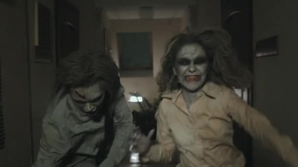 Tracking shot di inquietante zombie uomo e donna con trucco SFX e vestiti sporchi strappati che corrono lungo il corridoio buio - Filmati, video