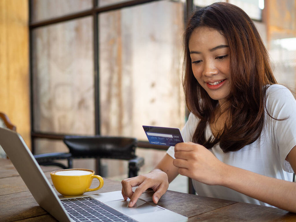 La joven asiática sonriente disfruta comprando en línea a través de una computadora y pagando en línea a través de tarjeta de crédito. Conveniencia de gastar sin efectivo. mantenerse a salvo, compras desde el hogar y la distancia social - Foto, imagen
