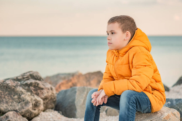 Pensive little boy in an orange jacket by the sea at sunset. Child's dreams. Rest and relaxation - Photo, Image